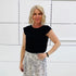 Woman wearing a black top and patterned skirt standing against a white wall.