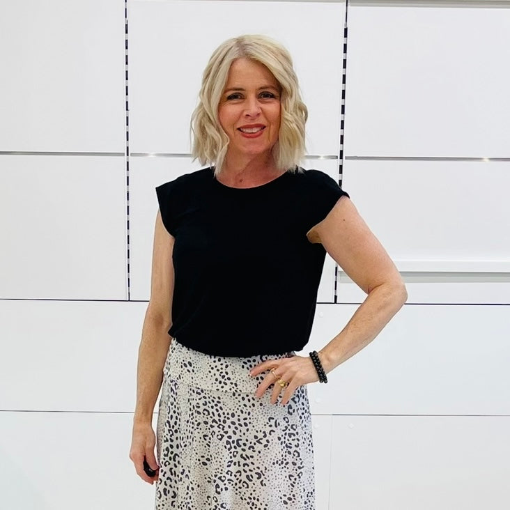 Woman wearing a black top and patterned skirt standing against a white wall.