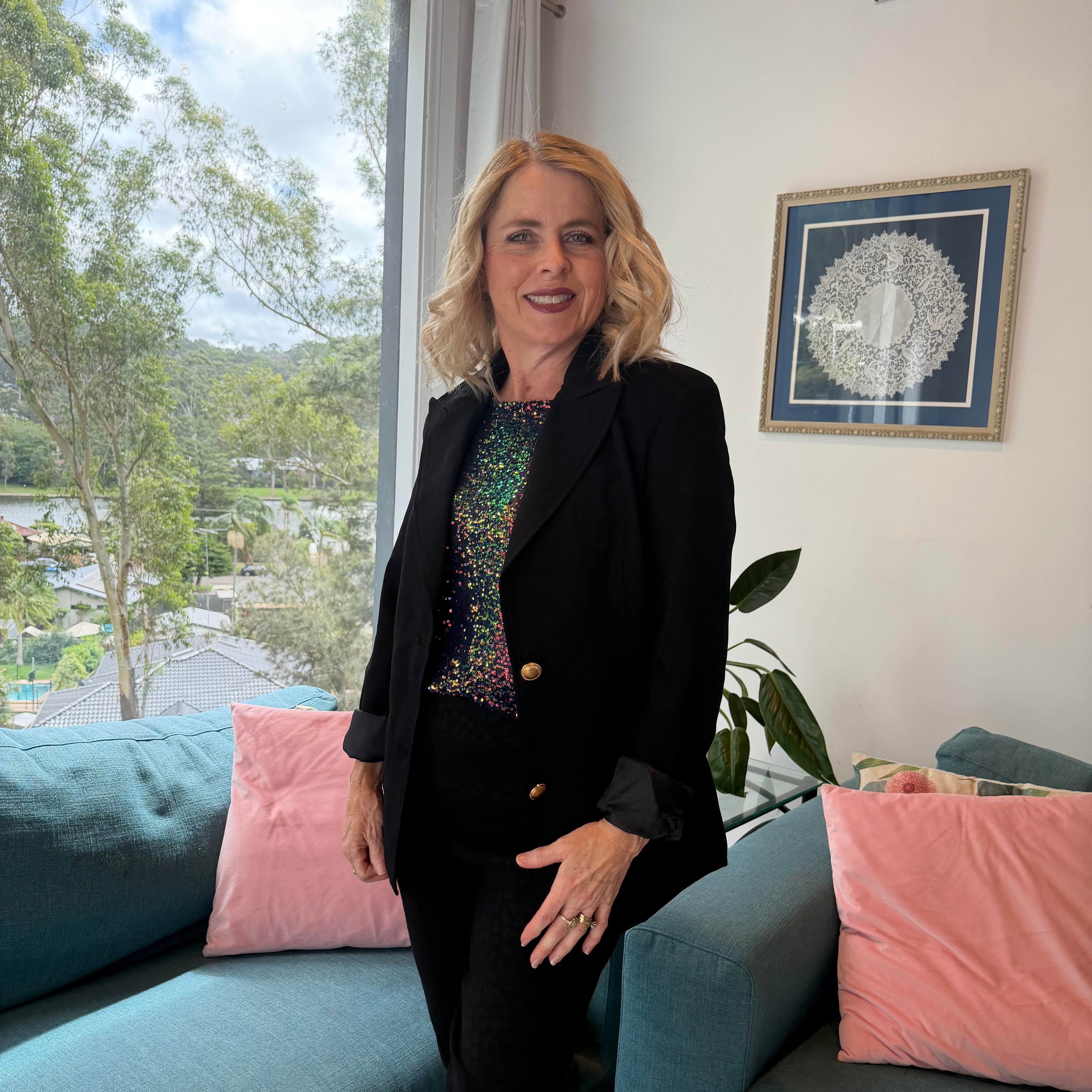 Woman standing in a room with a large window showing a scenic view, wearing a black blazer.