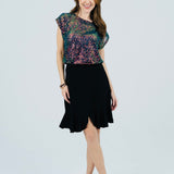 A woman modeling a two-piece outfit consisting of a black flared skirt and a sheer floral lace top with a dark green base and pink, purple, and blue floral patterns, paired with beige high-heeled sandals, against a light gray studio background.