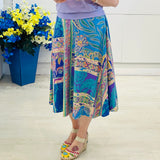 Woman wearing a purple top and colorful skirt standing next to a blue vase with flowers against a white wall.