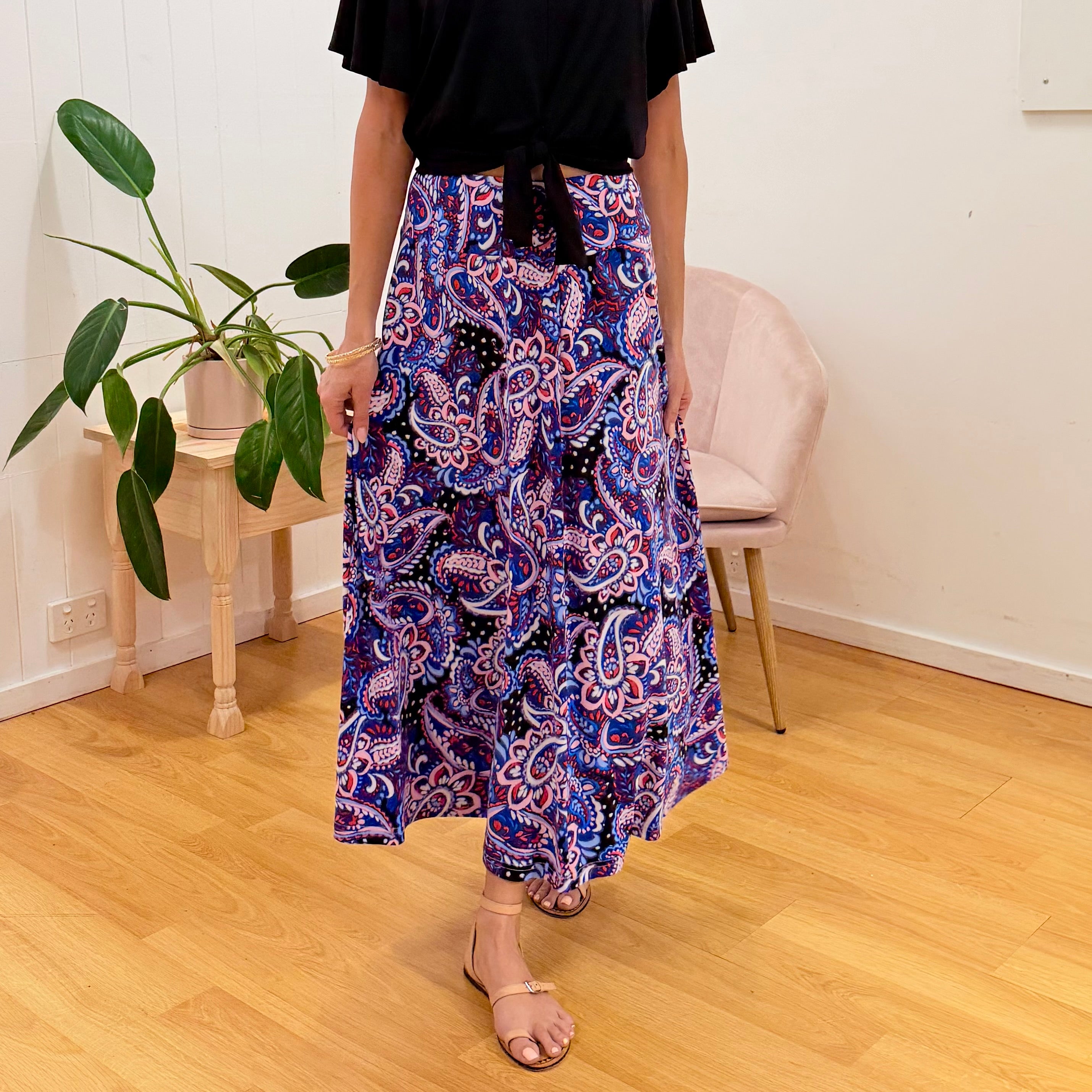 Person wearing a colorful patterned skirt in a room with a plant and chair.