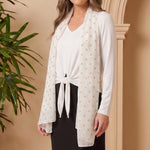 Woman in a white cardigan and black skirt standing next to a potted plant against a beige wall.