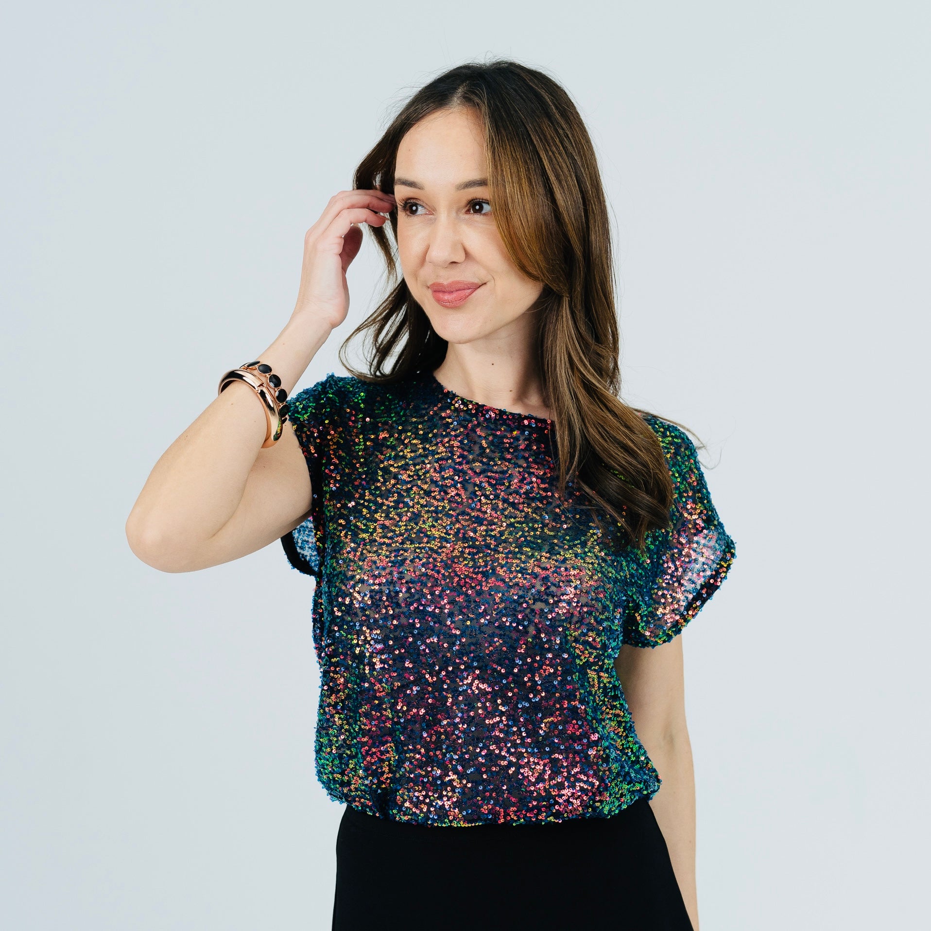 A woman modeling a short-sleeve crewneck top covered in multicolor sequins, paired with a black skirt, against a light gray studio background.