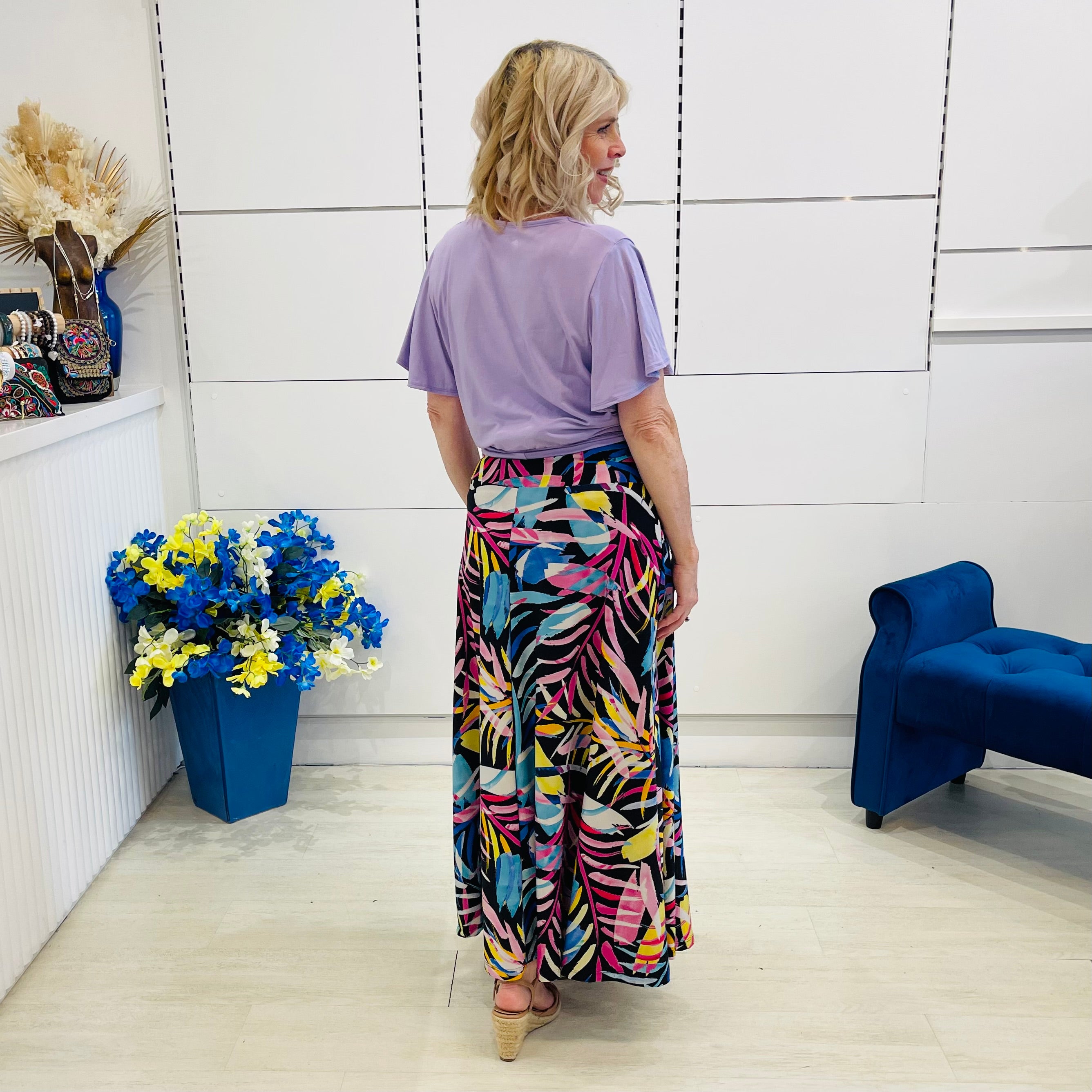 Woman wearing a colorful skirt and purple top in a room with a blue chair and floral arrangement.
