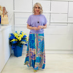 Woman wearing a purple top and colorful patterned skirt standing in a room with a white wall and a blue flower pot.