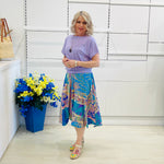 Woman wearing a purple top and colorful skirt standing next to a blue vase with flowers against a white wall.