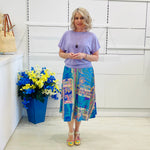 Woman wearing a purple top and colorful skirt standing next to a blue vase with flowers against a white wall.