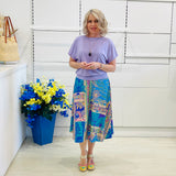 Woman wearing a purple top and colorful skirt standing next to a blue vase with flowers against a white wall.