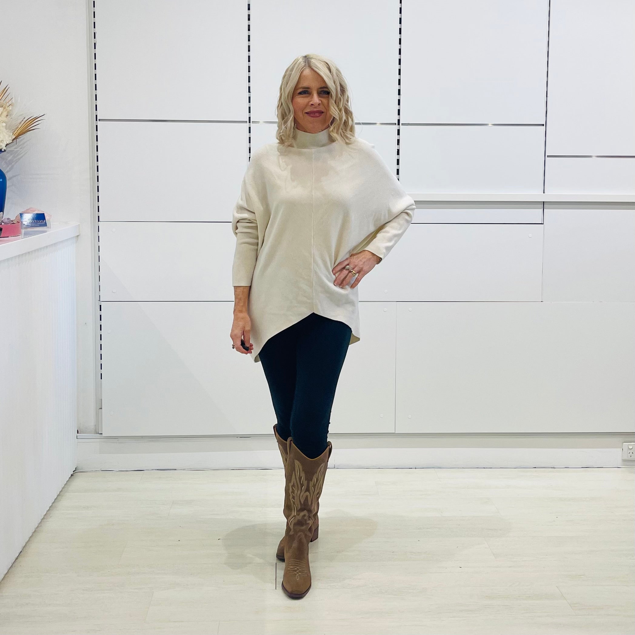 Cream knit, long sleeve, high neckline, asymmetrical hemline, winter 2025 jumper sweater. With black pants and and brown boots with embroidery.