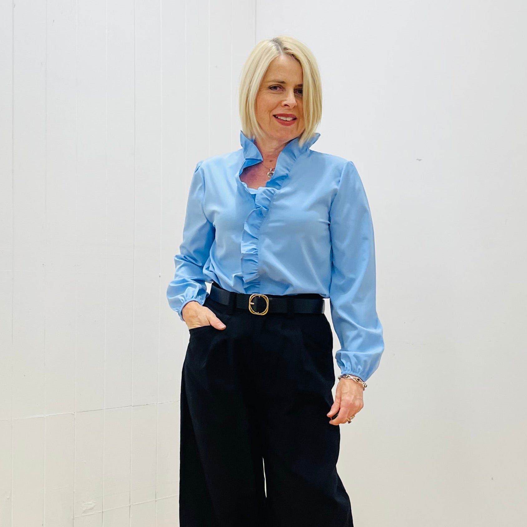 Woman wearing a light blue shirt and black pants standing in a room with white walls and wooden floor.