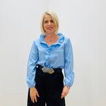 Woman wearing a light blue blouse with ruffled collar and black pants against a white background