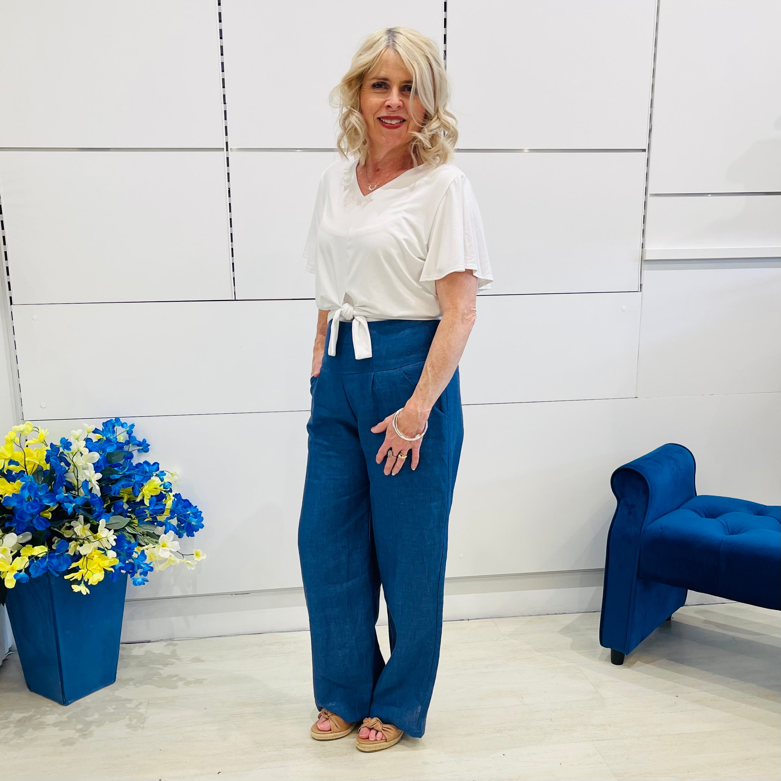 Woman in a white top and blue pants standing in a room with a blue chair and flower arrangement.