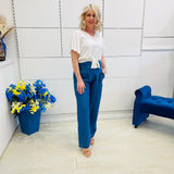 Woman in a white top and blue pants standing in a room with a blue chair and flower arrangement.
