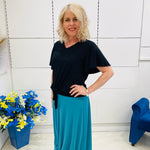 Woman in black top and blue pants standing in a room with flowers and a chair.