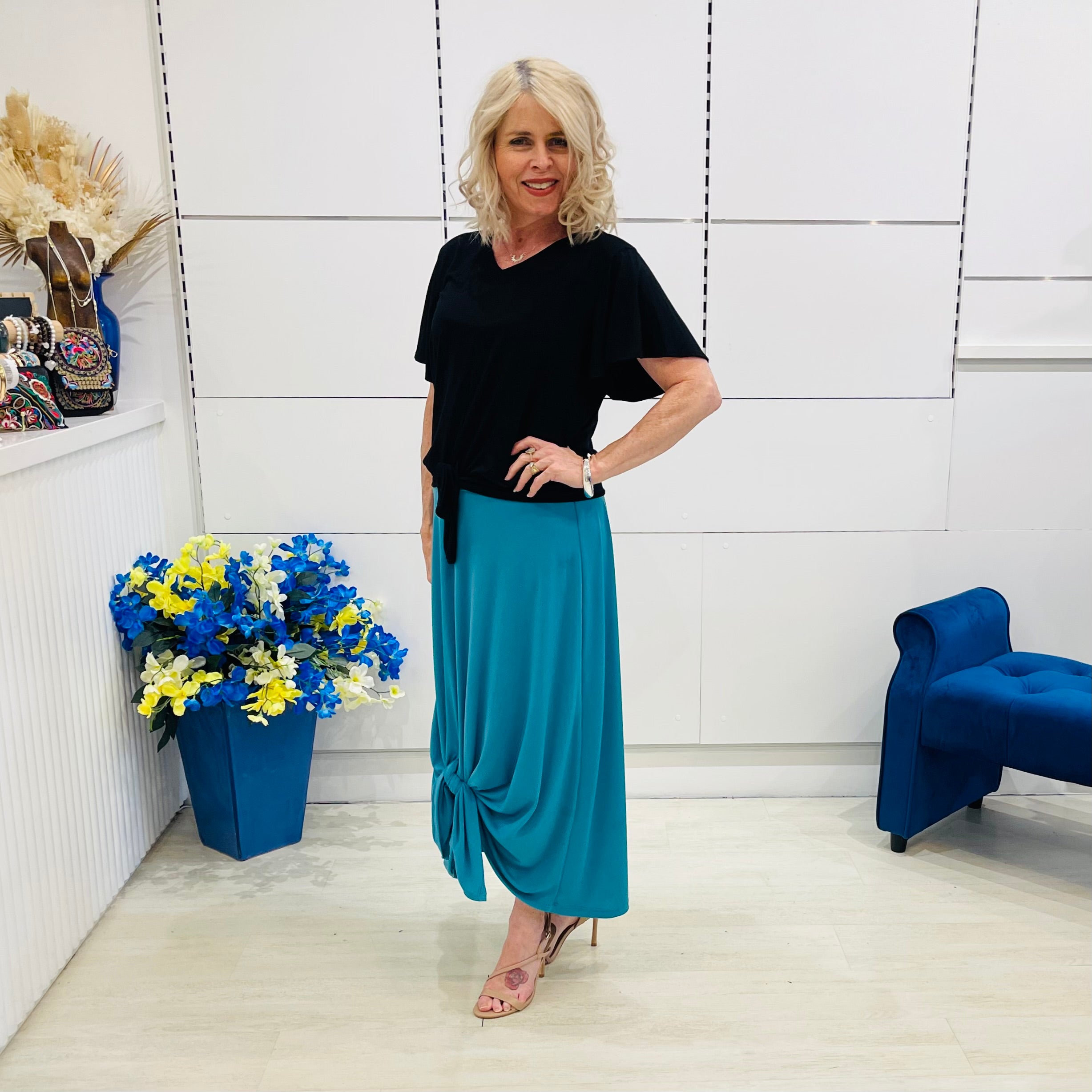 Woman in black top and blue skirt standing in a room with white walls, blue chair, and flower arrangement.