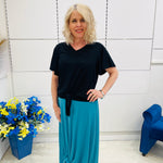 Woman wearing a black top and blue skirt standing in a room with a white wall and blue chair.
