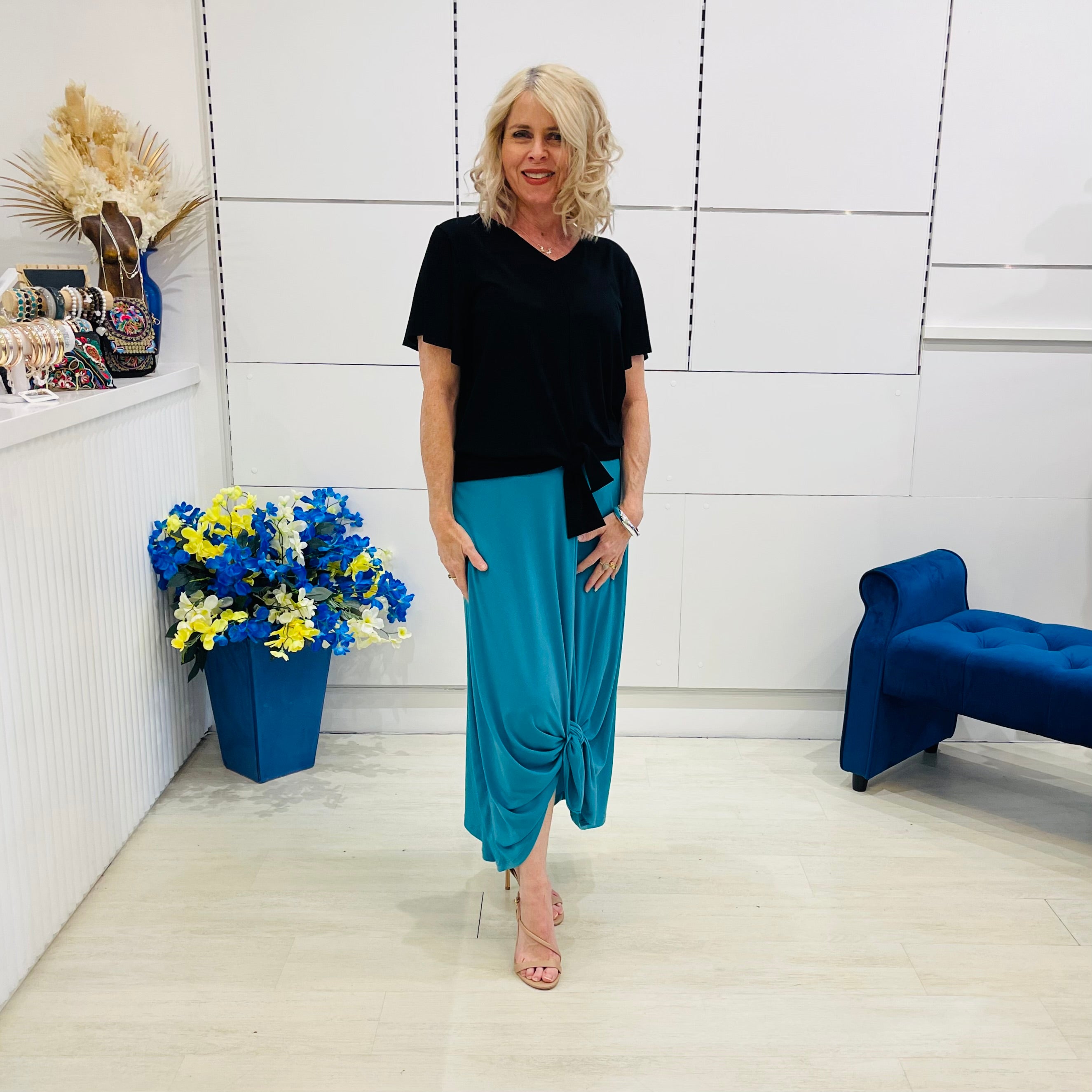 Woman in black top and blue pants standing in a room with white walls, a blue chair, and a flower arrangement.