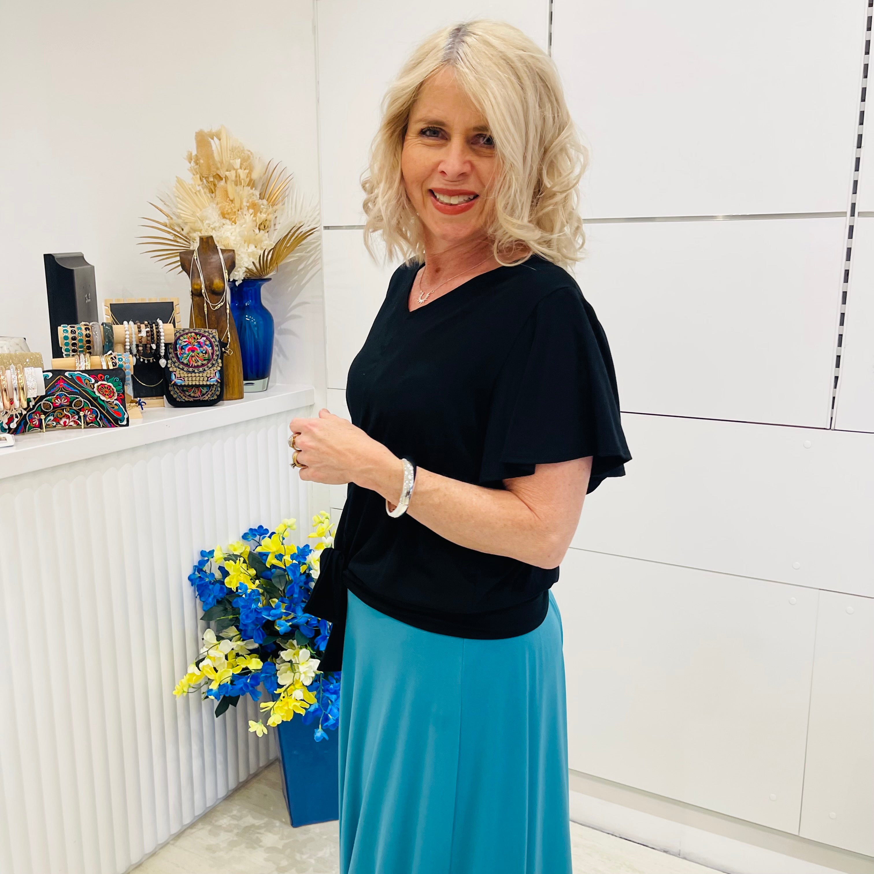 Woman in a black top and blue skirt standing in a room with decorative items on a shelf.