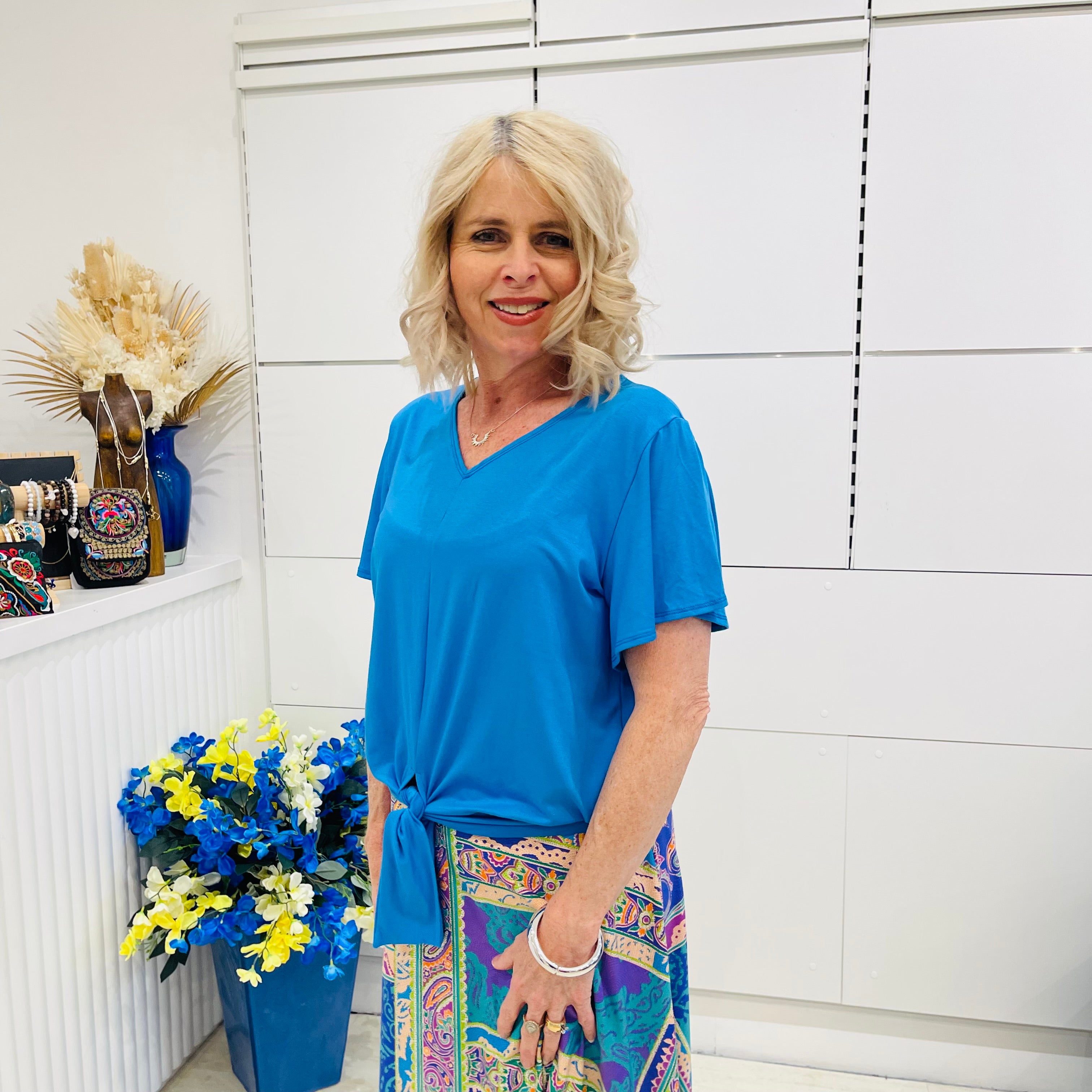 Woman in a blue top and colorful skirt standing in front of a white tiled wall with decorative elements.