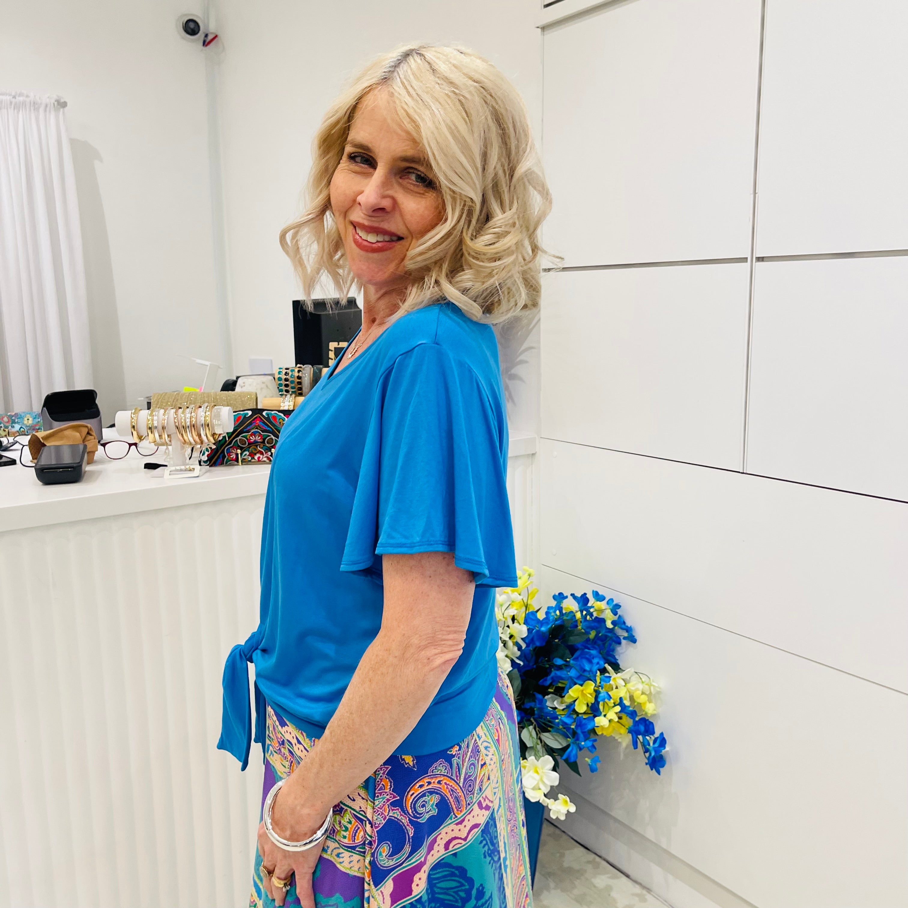 Woman in a blue top and colorful pants standing in a room with white walls and a radiator.