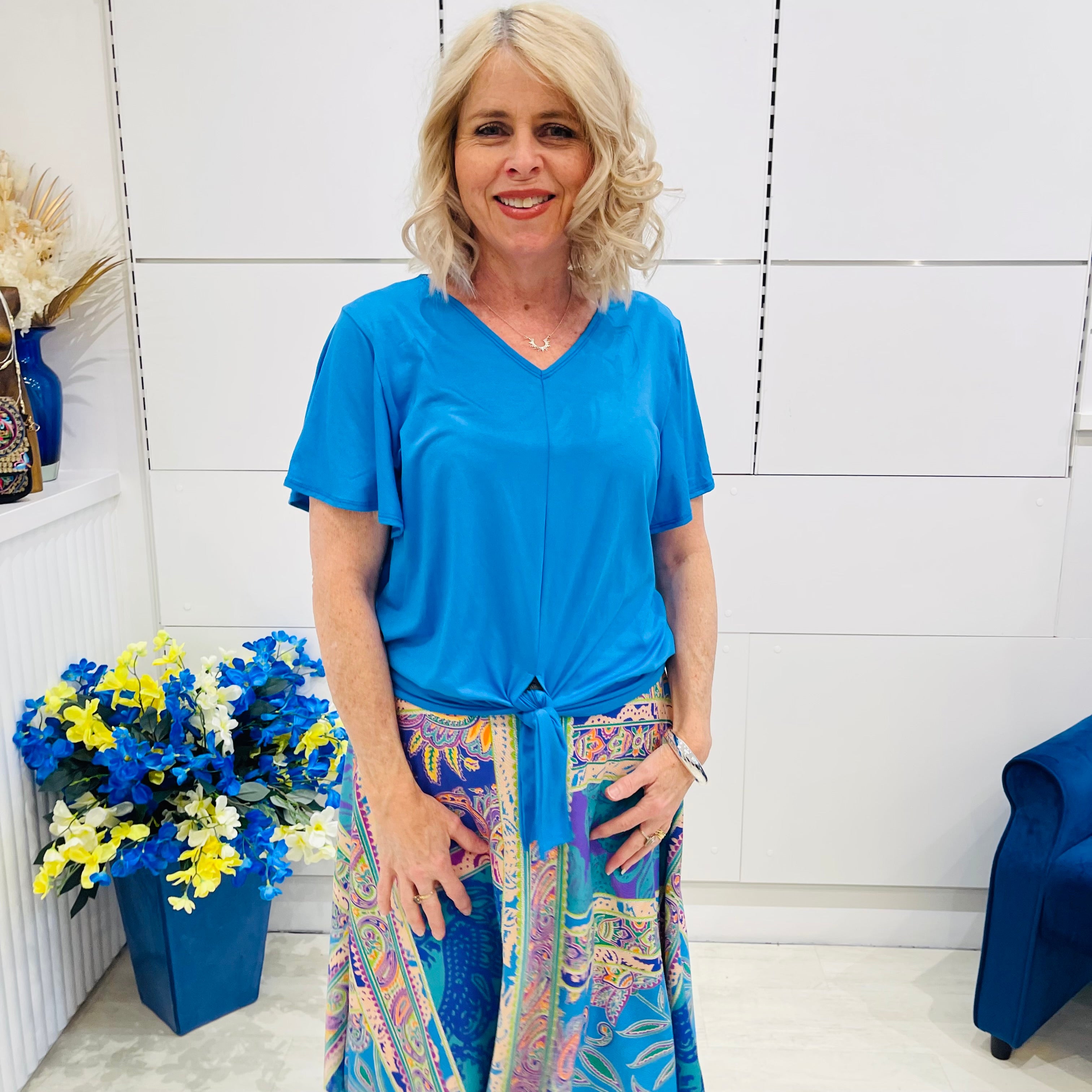 Woman wearing a blue top and colorful skirt standing in a room with white walls and decorative elements.