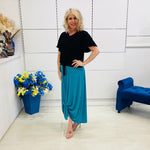 Woman in black top and teal skirt standing in a room with a blue chair and flower arrangement.