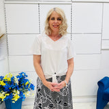 Woman in a white shirt and patterned skirt standing in front of a white wall with flowers and a blue ribbon.