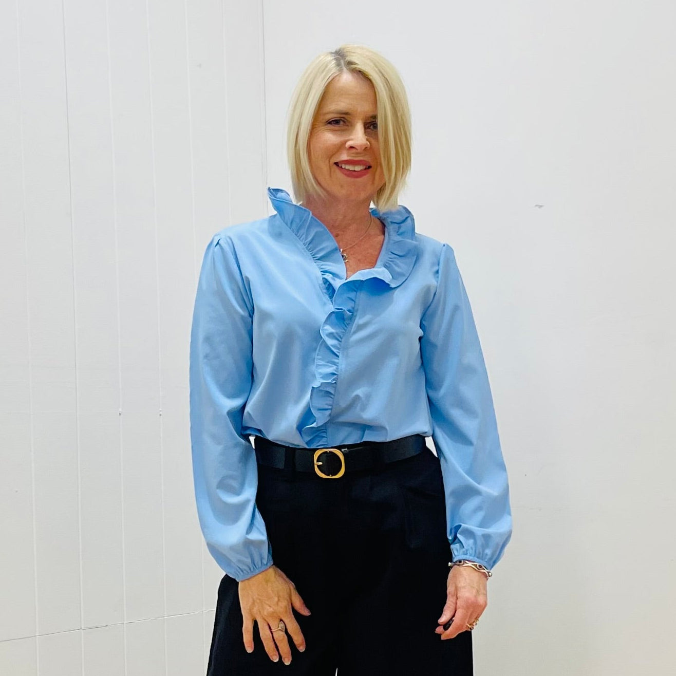 A woman wearing a pale blue long-sleeved shirt with gathered sleeves and a ruffle along the hem and center front, paired with black wide-leg trousers and black shoes.