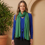 Woman wearing a blue top and green scarf standing next to a potted plant against a beige wall.