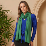 Woman wearing a blue cardigan and black skirt with a green scarf, standing in front of a beige wall with a plant.