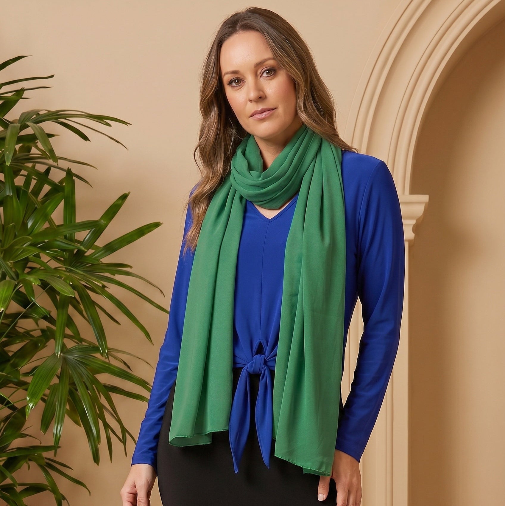 Woman wearing a blue cardigan and black skirt with a green scarf, standing in front of a beige wall with a plant.
