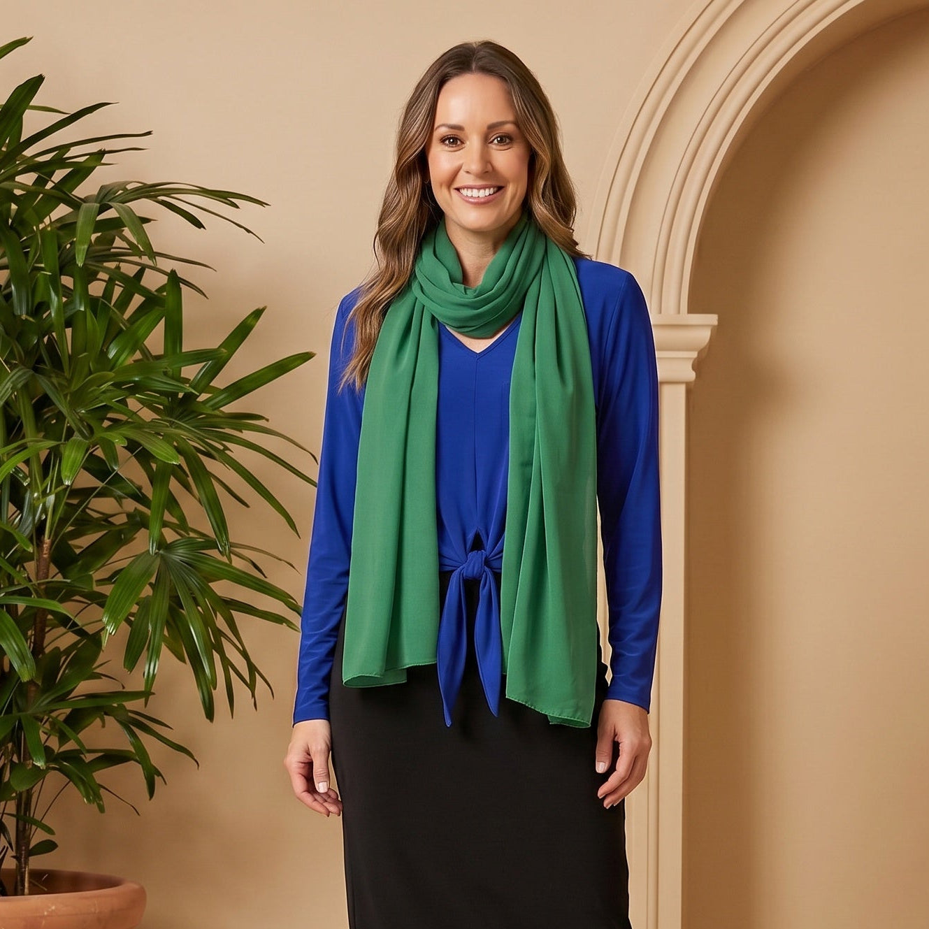 Woman wearing a blue top, green scarf, black skirt, and brown boots standing next to a potted plant against a beige wall.