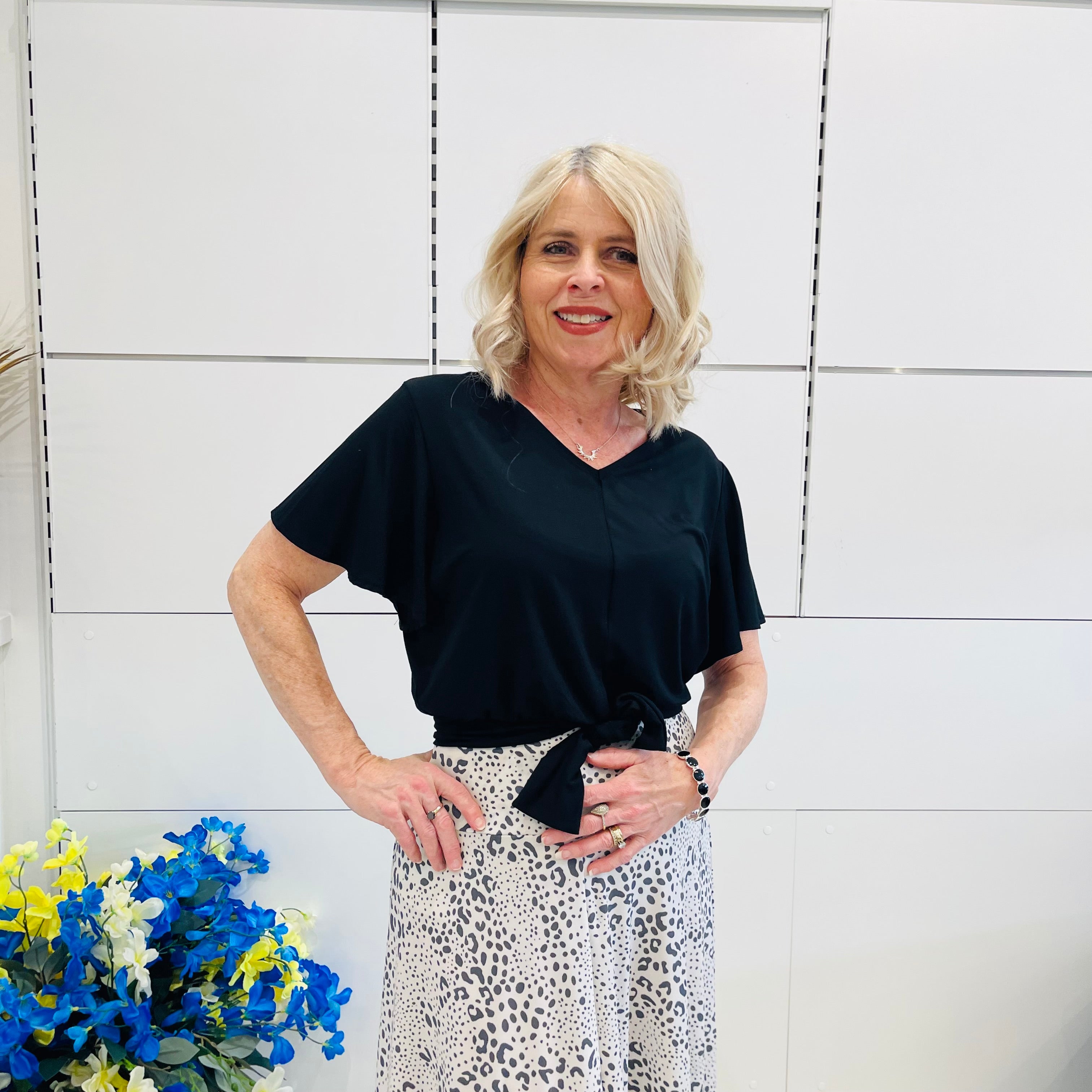 Woman wearing a black top and patterned skirt standing against a white tiled wall with flowers in the foreground.