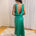 A woman stands with her back to the camera wearing a sleeveless emerald green maxi dress with a deep open back, standing on a patterned rug in front of white curtains.