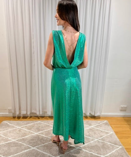 A woman stands with her back to the camera wearing a shimmering emerald green sleeveless maxi dress with a deep V-back. She is standing on a gray geometric patterned rug in front of white curtains and a light wood floor.