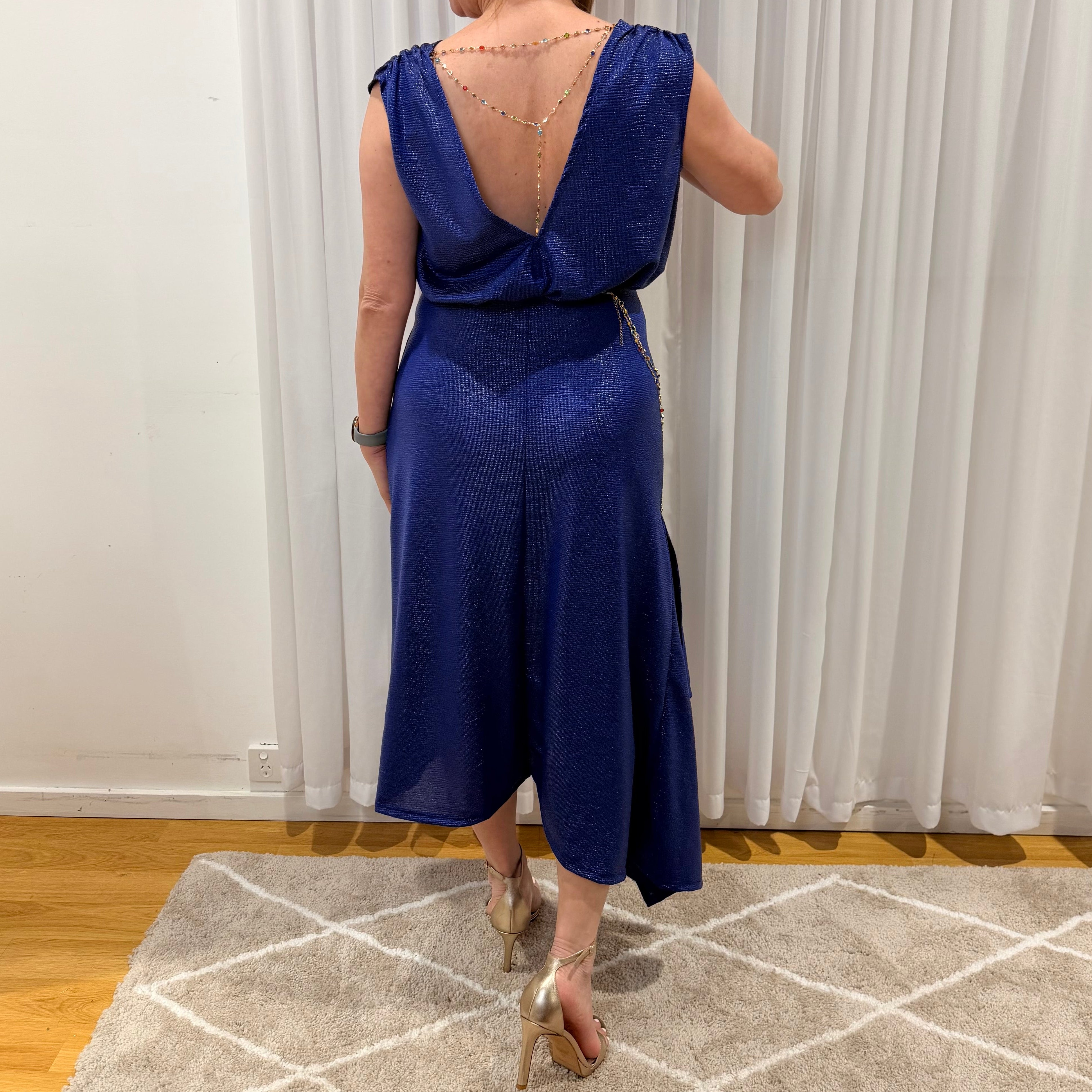 A woman wearing a royal blue sleeveless wrap-style midi dress with a deep V-back and tie detail at the waist, shown from behind in an indoor setting with white curtains and a patterned rug.