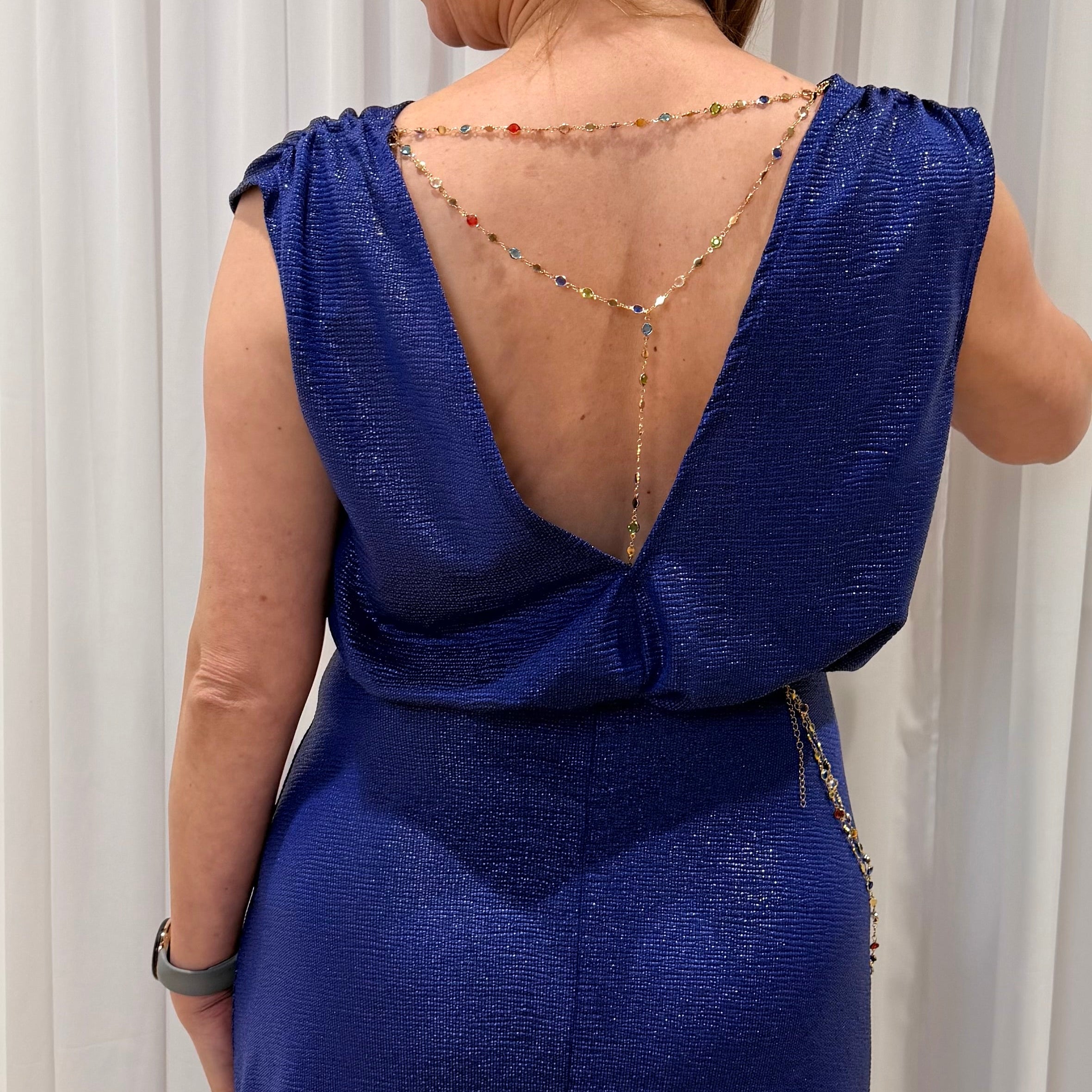A woman shown from the back wearing a metallic royal blue sleeveless dress with a deep open back, decorated with a delicate beaded gold chain draped across her upper back.