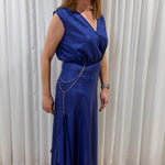 A woman wearing a sleeveless royal blue wrap-style maxi dress with a draped chain detail at the waist, standing on a patterned rug in front of white curtains.