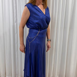 A woman wearing a sleeveless royal blue wrap-style maxi dress with a draped chain detail at the waist, standing on a patterned rug in front of white curtains.
