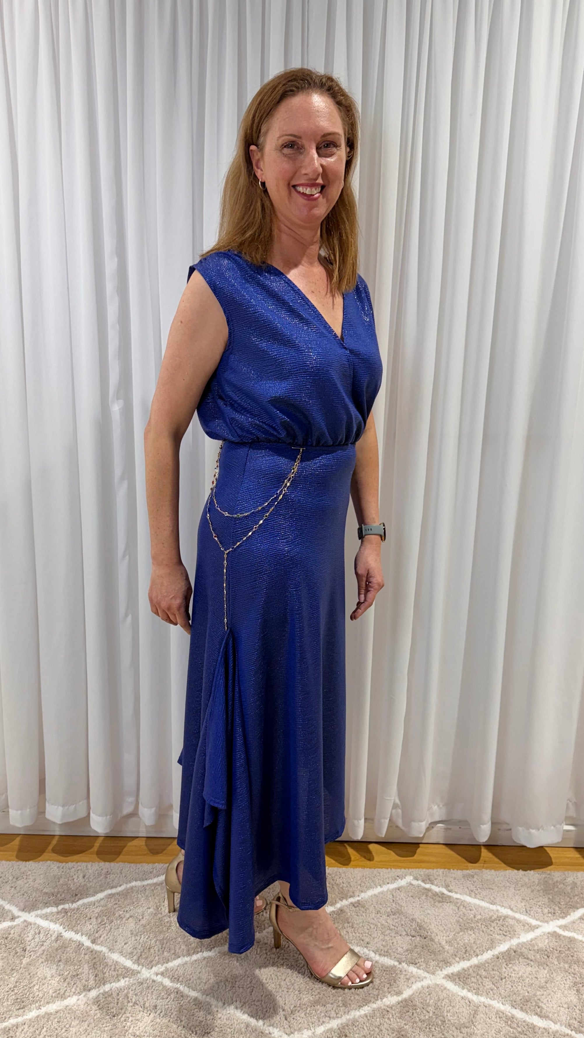 A woman wearing a sleeveless royal blue wrap-style maxi dress with a draped chain detail at the waist, standing on a patterned rug in front of white curtains.