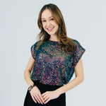 A woman modeling a short-sleeve top covered in multicolored sequins, paired with a black skirt, against a light gray studio background.