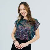 A woman modeling a short-sleeve top covered in multicolored sequins, paired with a black skirt, against a light gray studio background.