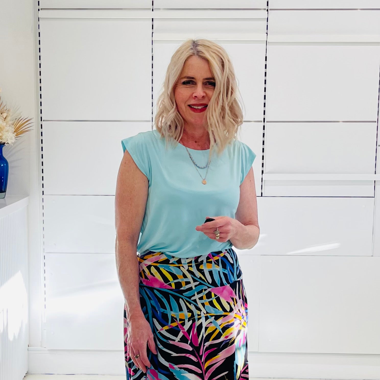 Woman wearing a light green top and colorful skirt standing against a white wall.