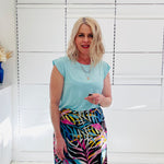 Woman wearing a light green top and colorful skirt standing against a white wall.