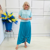 Woman wearing a light blue top and teal skirt standing in a room with a white wall and a decorative plant.