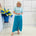 Woman wearing a light blue top and teal skirt standing in a room with a white wall and a decorative plant.