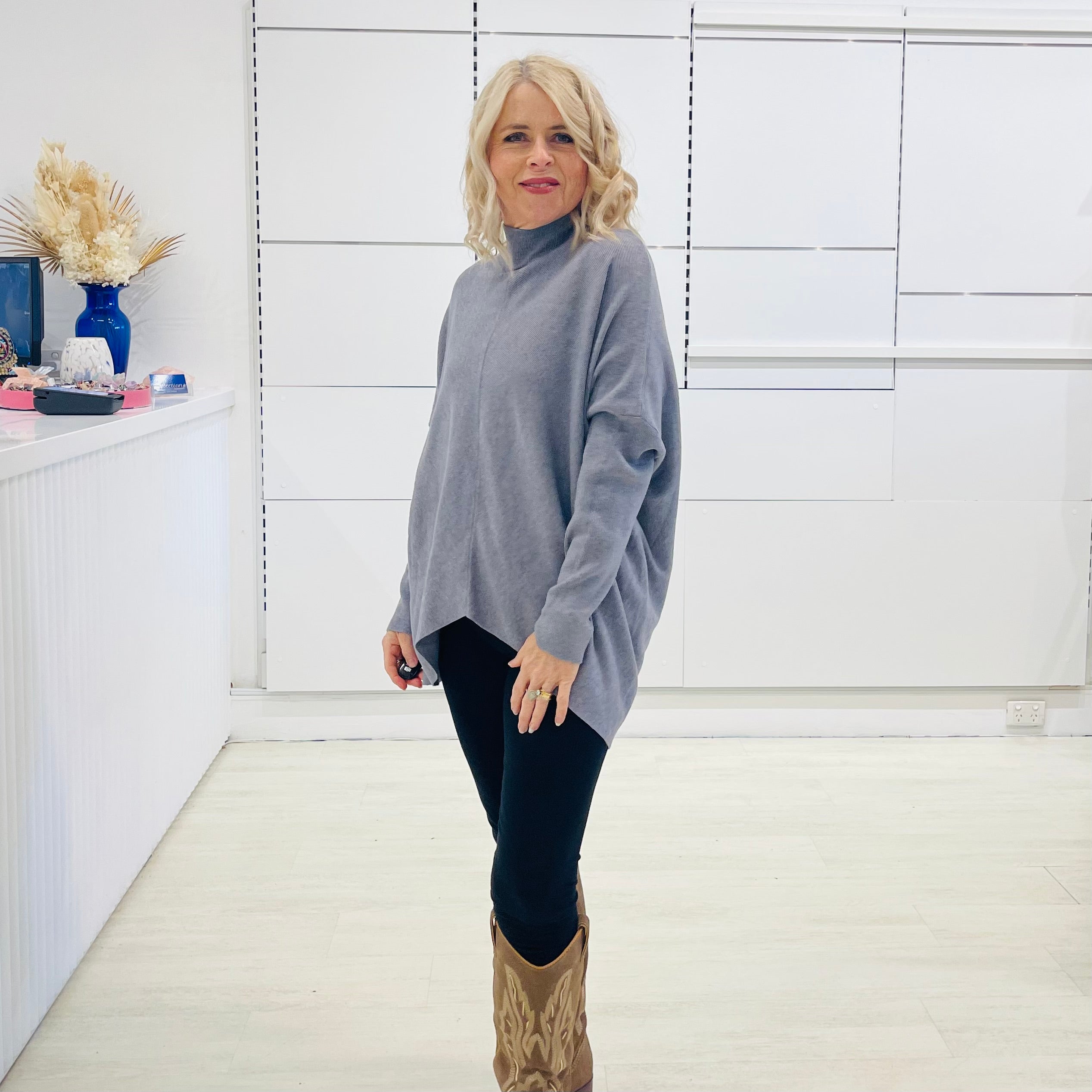 Grey knit sweater with long sleeves, high neckline and asymmetrical hemline. With black pants and brown embroidered boots.