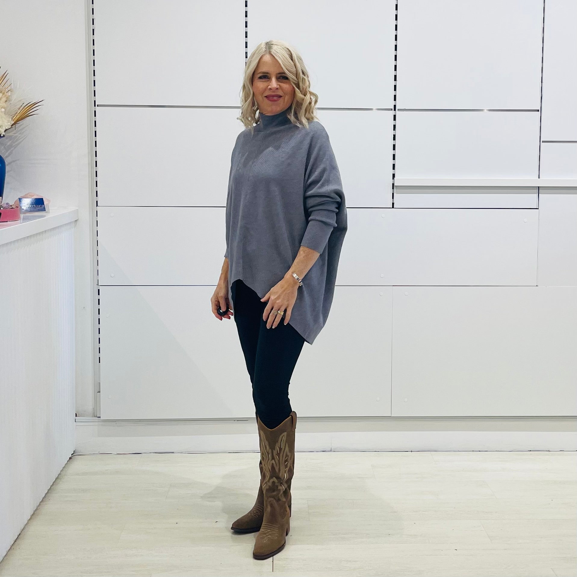 Grey knit sweater with long sleeves, high neckline and asymmetrical hemline. With black pants and brown embroidered boots.