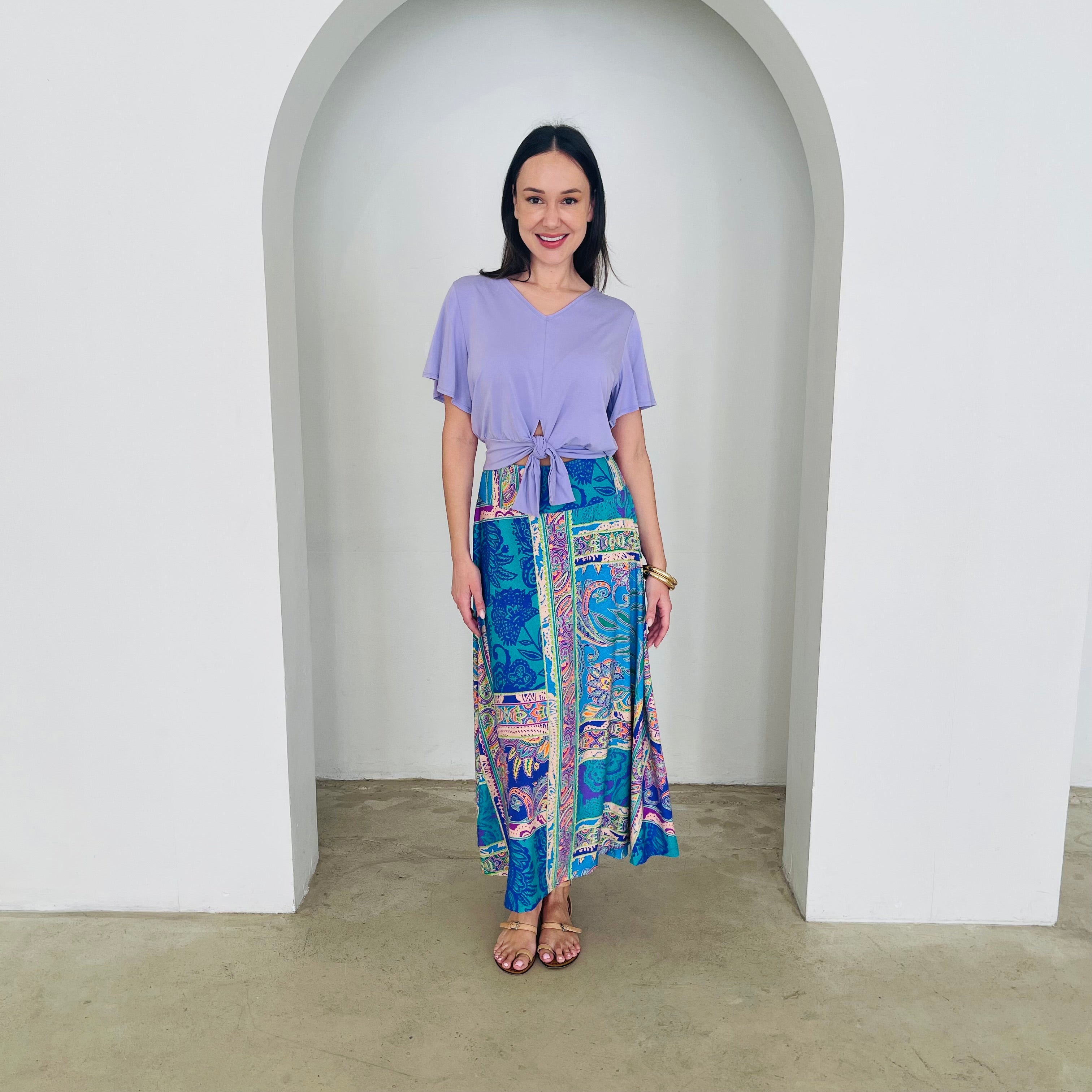 Woman wearing a purple top and colorful patterned skirt standing in front of a white archway.
